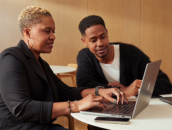 Twee Apple medewerkers zitten samen aan een tafel op een MacBook te werken. 