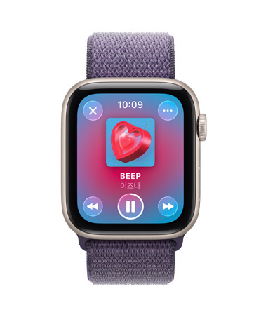 미디어 재생 기능에 Apple Music이 표시되고 오른쪽 옆면에 Digital Crown이 보이는 스타라이트 색상 알루미늄 케이스의 Apple Watch SE 3, 그리고 퍼플 포그 색상 스포츠 루프