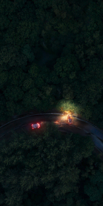 An overheard view of a winding road at night, two vehicles perpendicular with headlines on, surrounded by trees