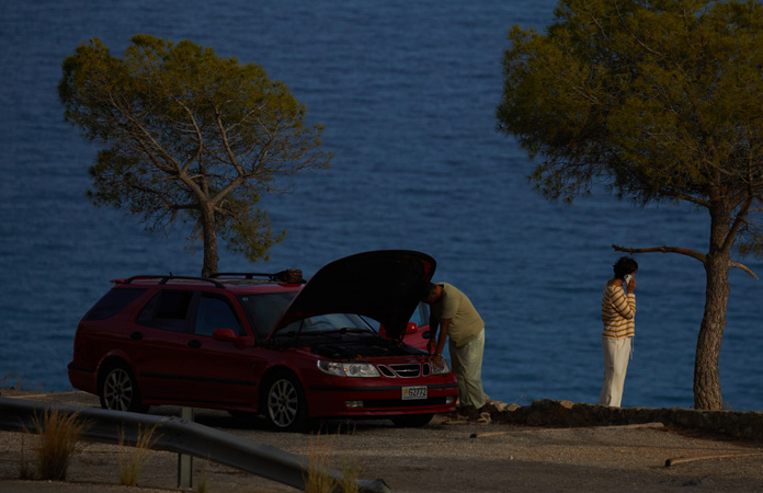 A car stranded by the side of a road with one person by the car and one person using iPhone to contact roadside assistance