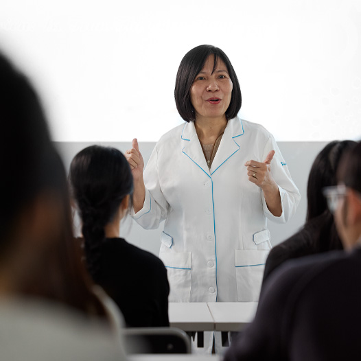 An instructor in a white lab coat teaches at the front of a classroom full of students